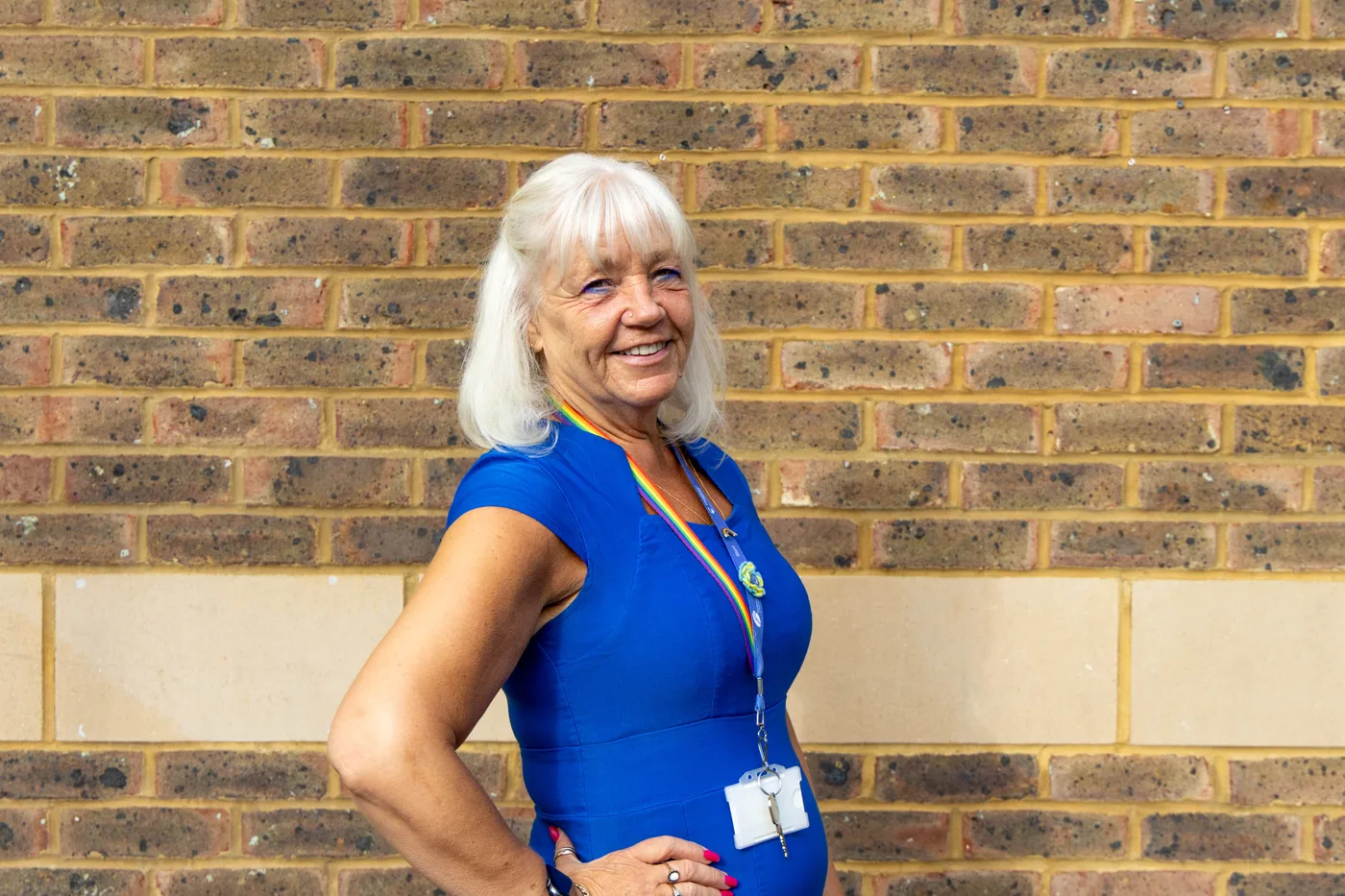 Michelle Chapman, in a blue dress and lanyard, standing with one hand on her hip against a brick wall